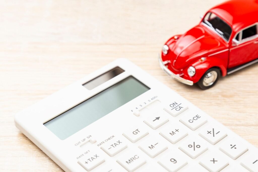 A close-up of a white calculator and a small red toy car on a light wooden surface. The image suggests concepts of car costs or budgeting.