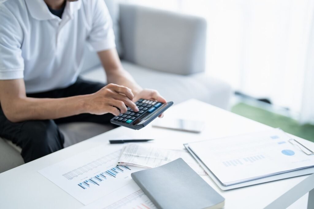 A person in a white shirt holds a calculator while sitting on a couch, surrounded by papers, financial graphs, and a notebook on a white table.