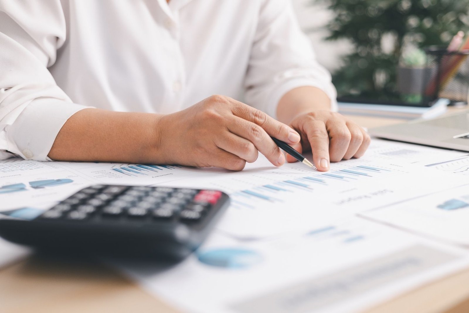 Close-up of hands analyzing financial charts and reports with pen, calculator, and documents on desk, representing budgeting, data analysis, accounting, and financial planning in office setting.