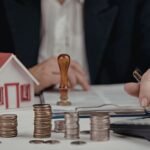 Person calculating property finances at desk with house model, stacked coins, calculator, and documents, representing real estate investment, budgeting, mortgage planning, and financial analysis.